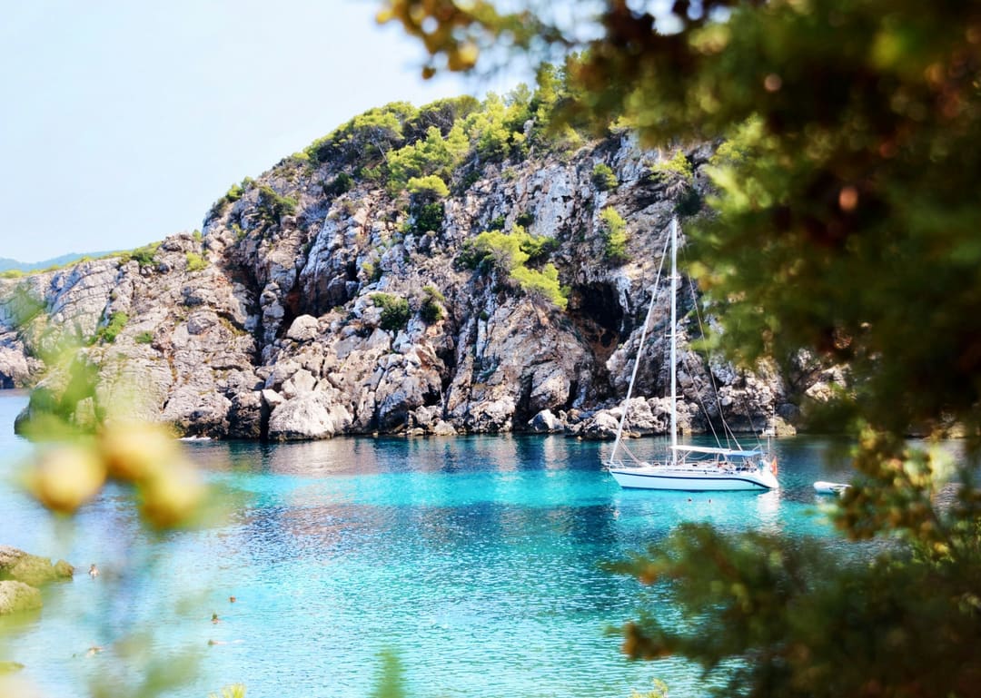 Velero fondeado en una cala de roca y pinos, con agua turquesa.